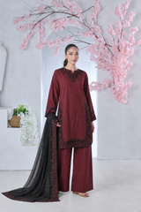 Woman in a maroon embroidered tunic and wide-leg trousers holding a black fabric shawl in front of pink blossoms.