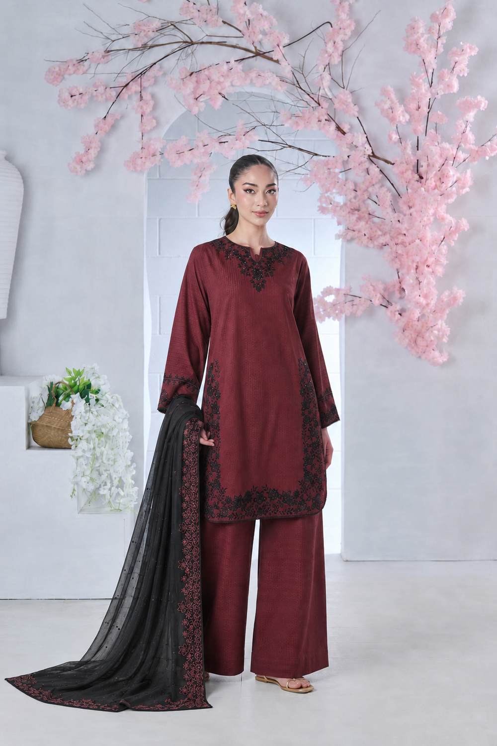 Woman in a maroon embroidered tunic and wide-leg trousers holding a black fabric shawl in front of pink blossoms.